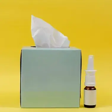 Box of tissues beside a nasal spray bottle against a yellow background