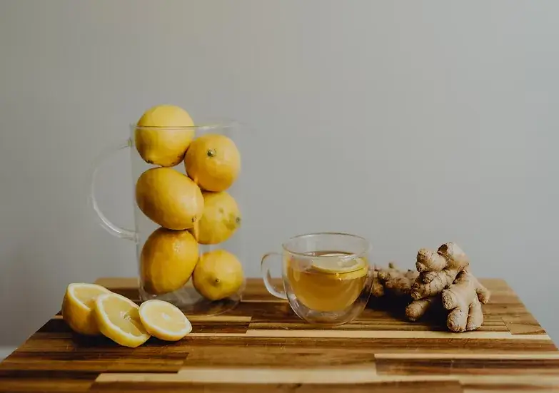 Glass cup of lemon and ginger tea beside fresh ginger root, lemon slices and a jug of whole lemons