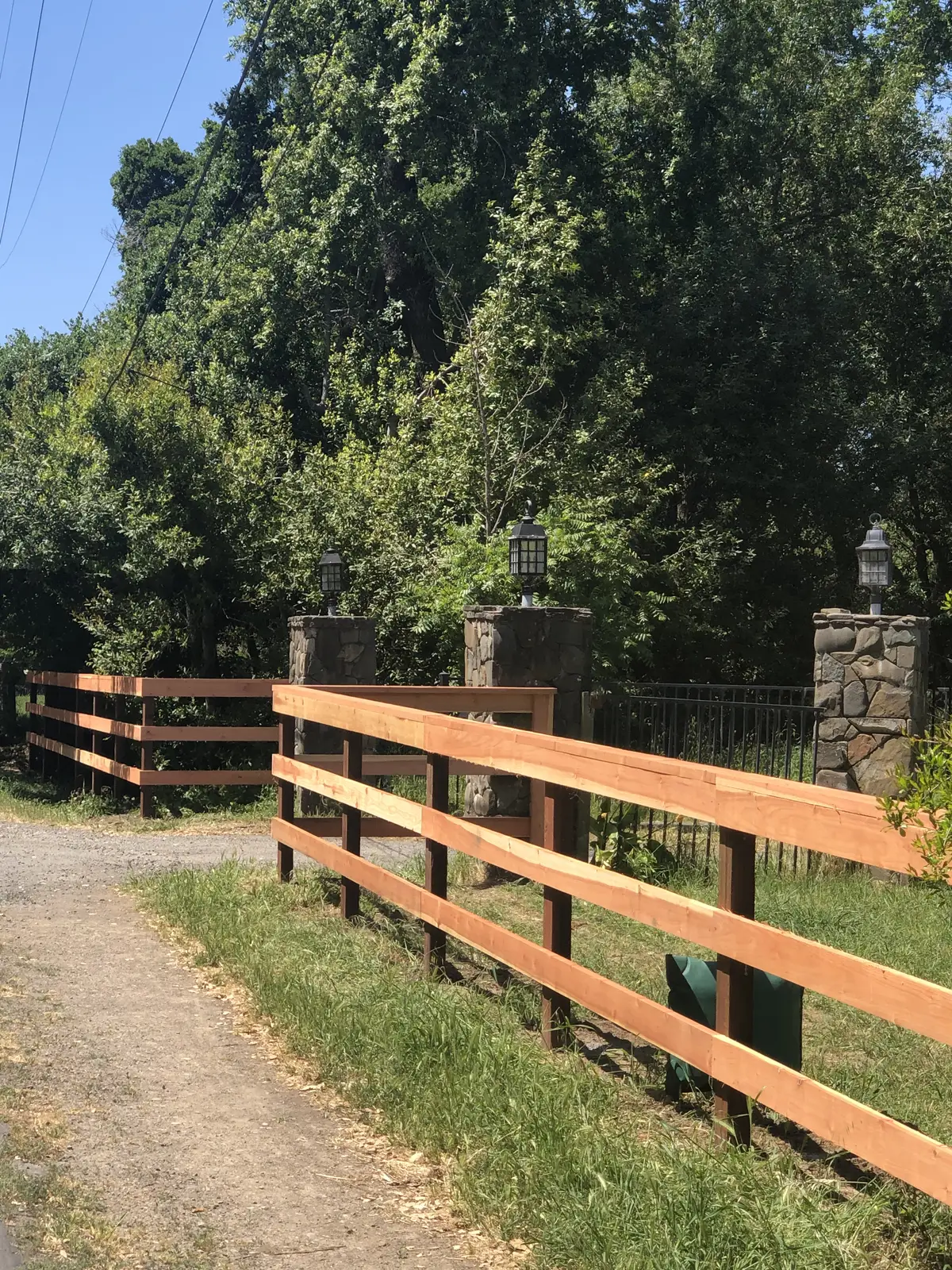 Three-rail redwood ranch fence at a driveway entrance flanked by stone pillars with lanterns