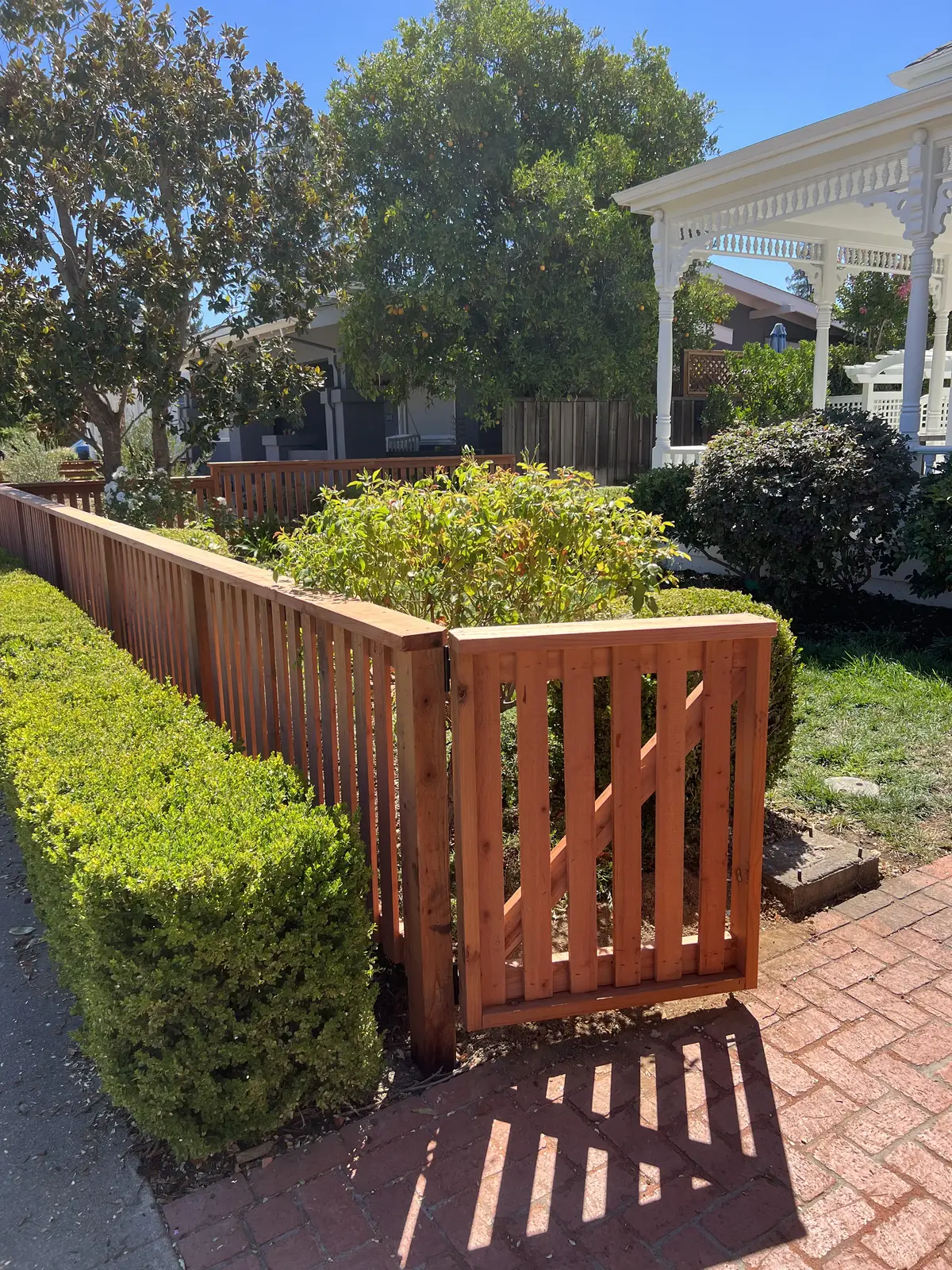 Redwood picket walk gate and matching fence beside a boxwood hedge on a brick path, white Victorian gazebo behind