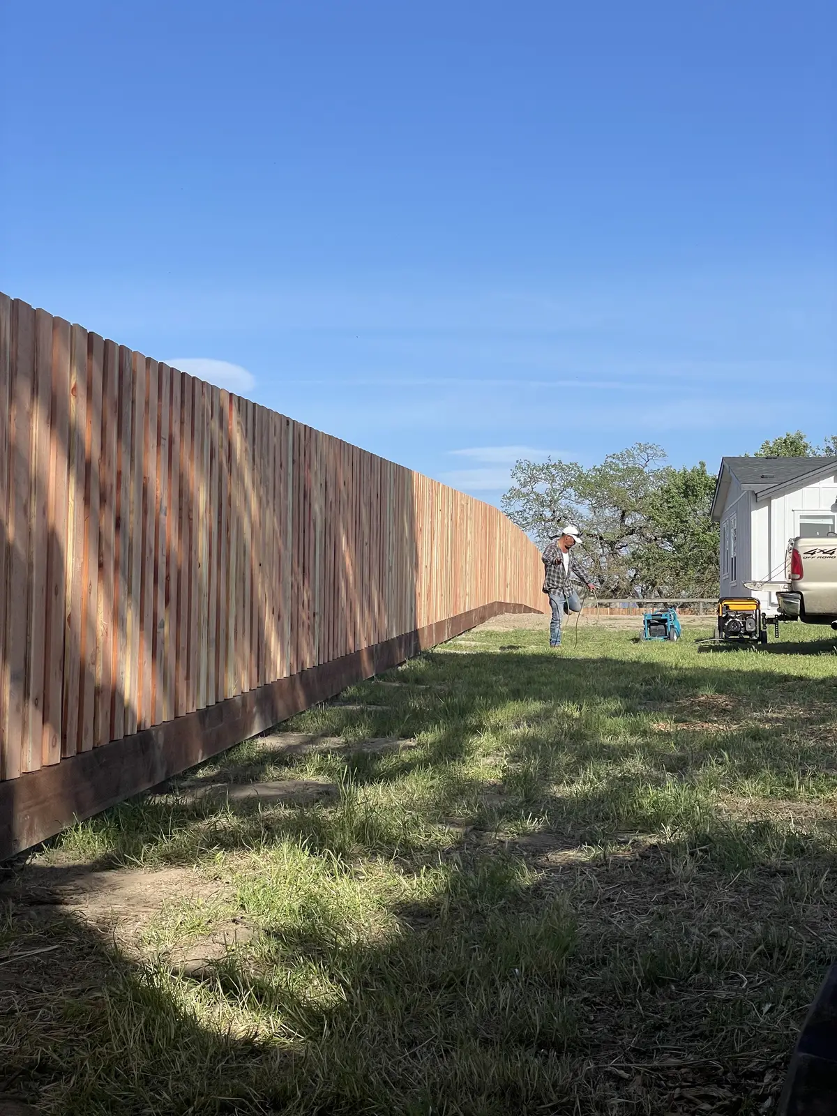 Long redwood board privacy fence with kickboard across a sunny lawn, crew member working at the far end
