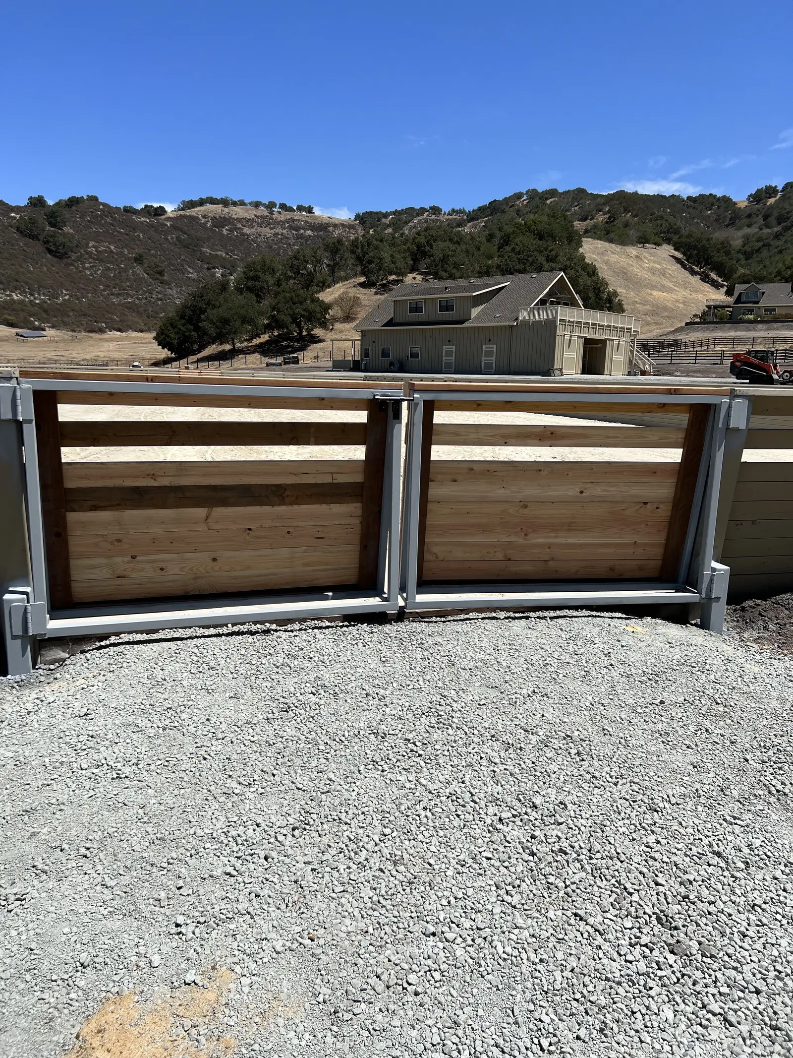 Closed double driveway gate with galvanized steel frames and horizontal redwood infill on a gravel drive, riding arena, barn and oak hills behind