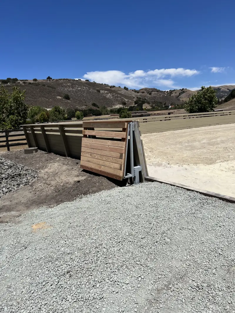 Welded steel-frame driveway gate with horizontal redwood infill swung open at the gravel entrance to a sand riding arena, horizontal board fence and dry hills behind