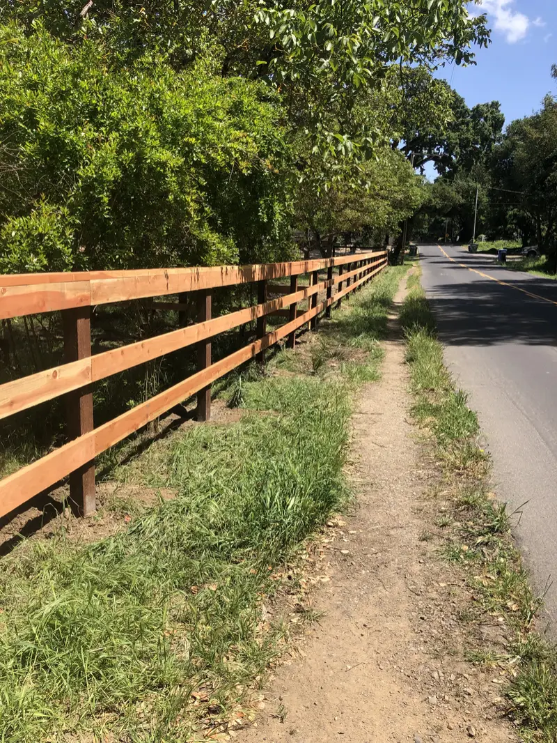 Three-rail rough-sawn redwood ranch fence on dark posts along a country road and dirt footpath