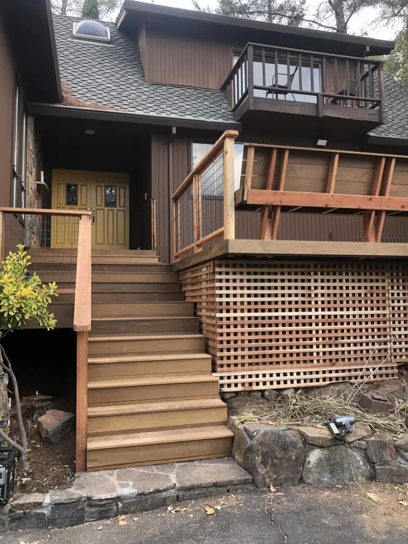 Redwood entry deck stairs with a welded-wire railing and a lattice skirt in front of a brown two-story house