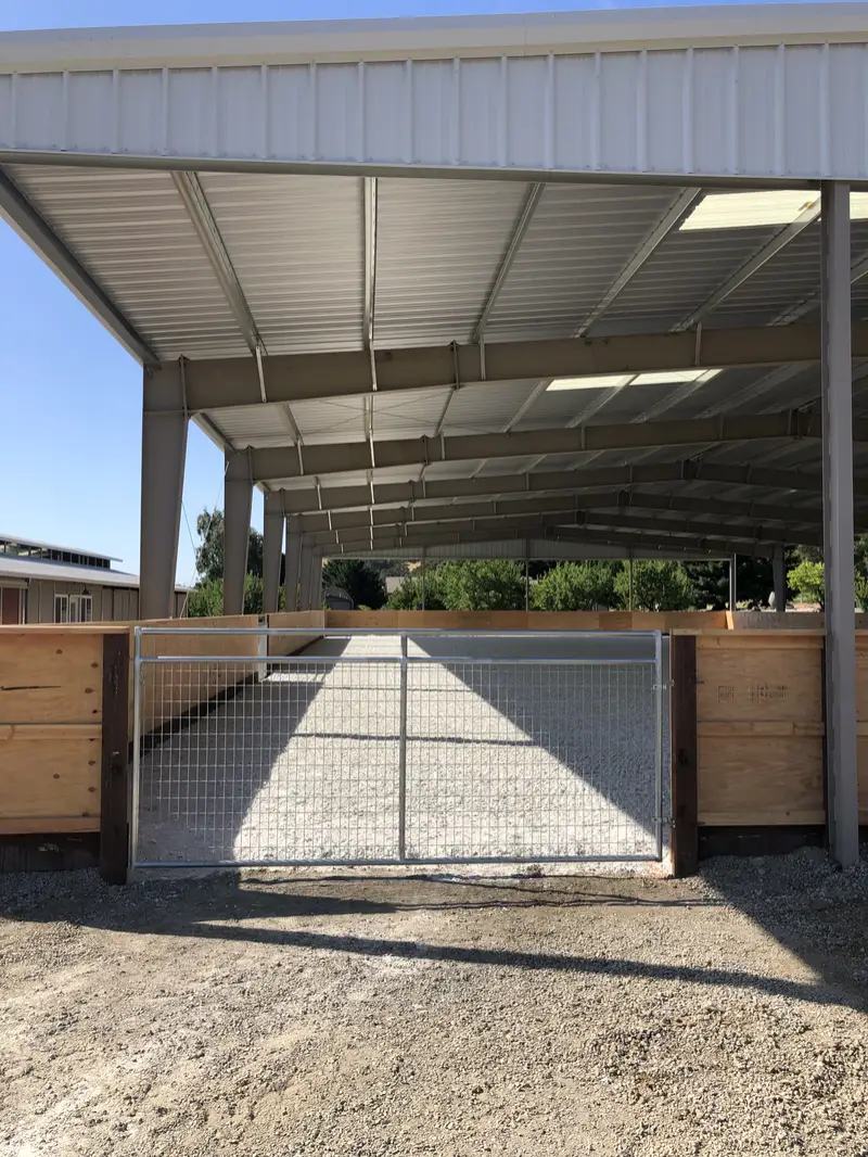 Covered riding arena with a solid wood kickwall and a galvanized wire-mesh ranch gate at the entrance
