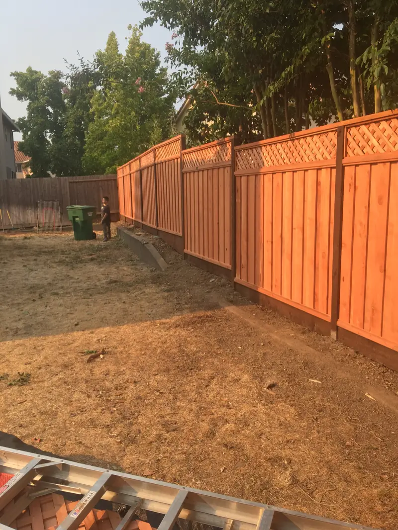 Long run of new redwood privacy fence with a lattice top and dark-stained posts across a dry backyard, seen from beside a work ladder