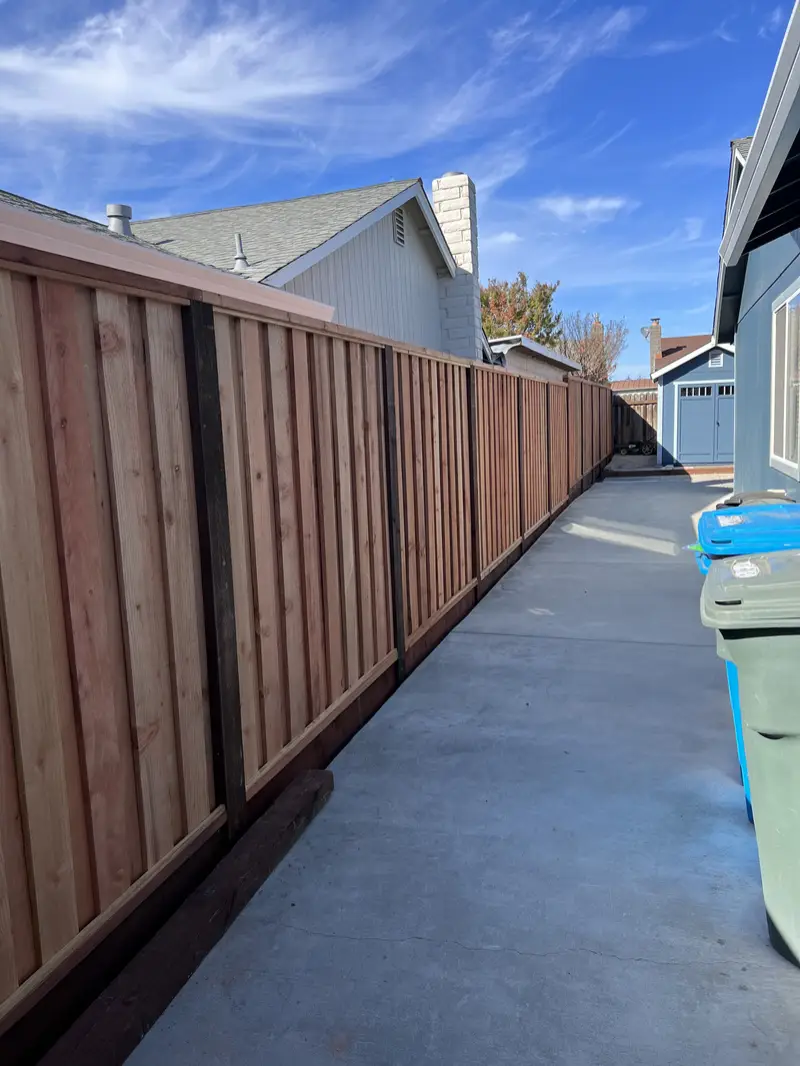 Long redwood board-and-batten privacy fence with dark-stained posts and kickboard running the length of a concrete side yard between homes