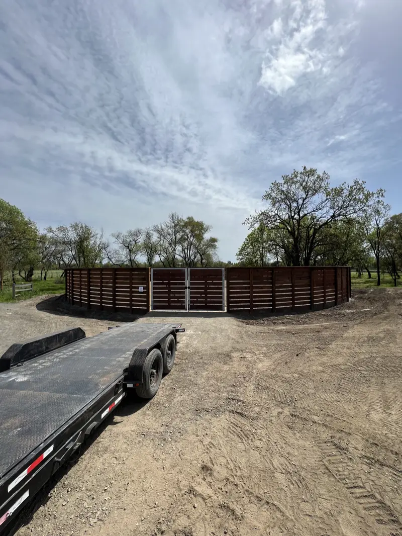 Solid horizontal redwood round pen with a steel-frame double gate on open oak land, flatbed trailer in the foreground