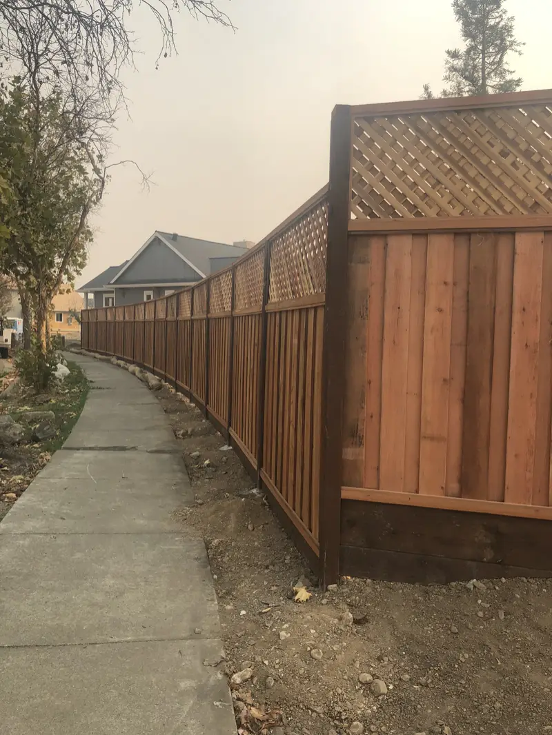 Lattice-top redwood privacy fence with dark-stained posts following a curving concrete path, hazy sky