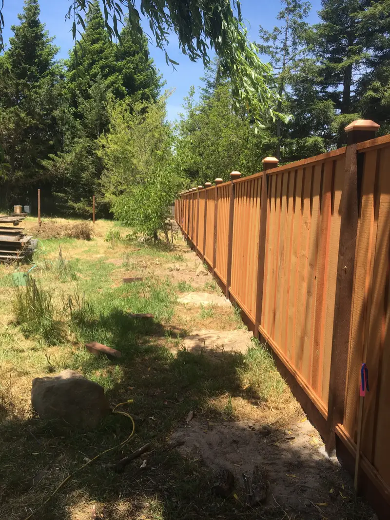 Long redwood board-and-batten fence with capped posts and a kickboard running along a wooded rural property line