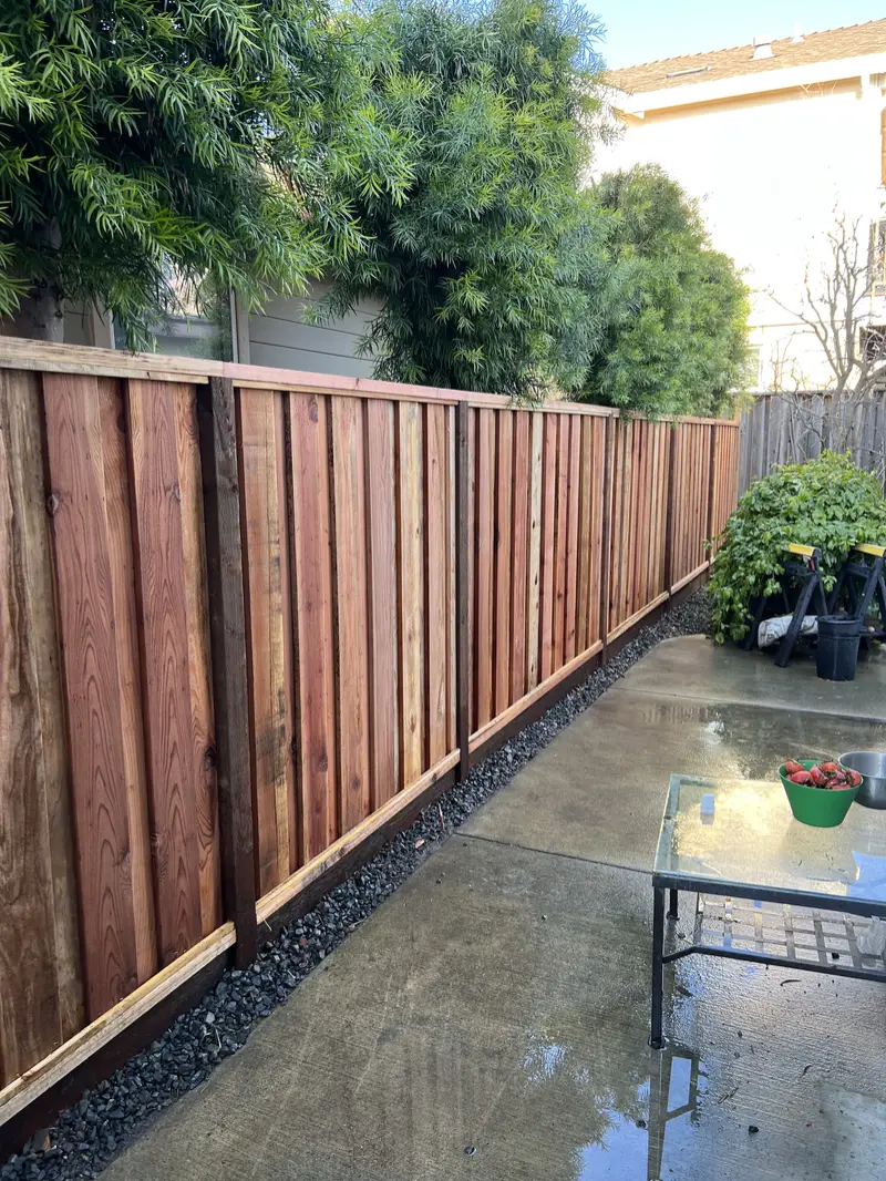 Redwood board-and-batten privacy fence with dark posts and kickboard along a concrete patio with a gravel strip