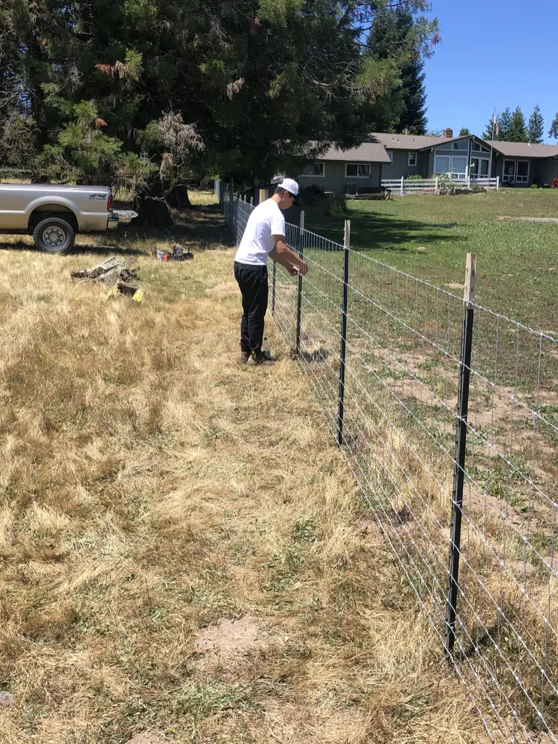 Crew member stretching woven-wire fencing onto steel T-posts in a dry field, pickup truck behind