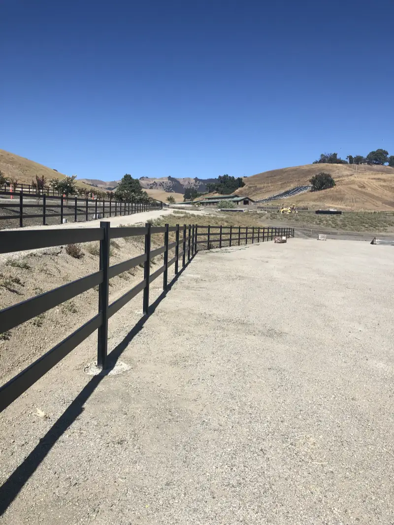 Black three-rail paddock fence along a gravel ranch lane with golden hills and oaks beyond
