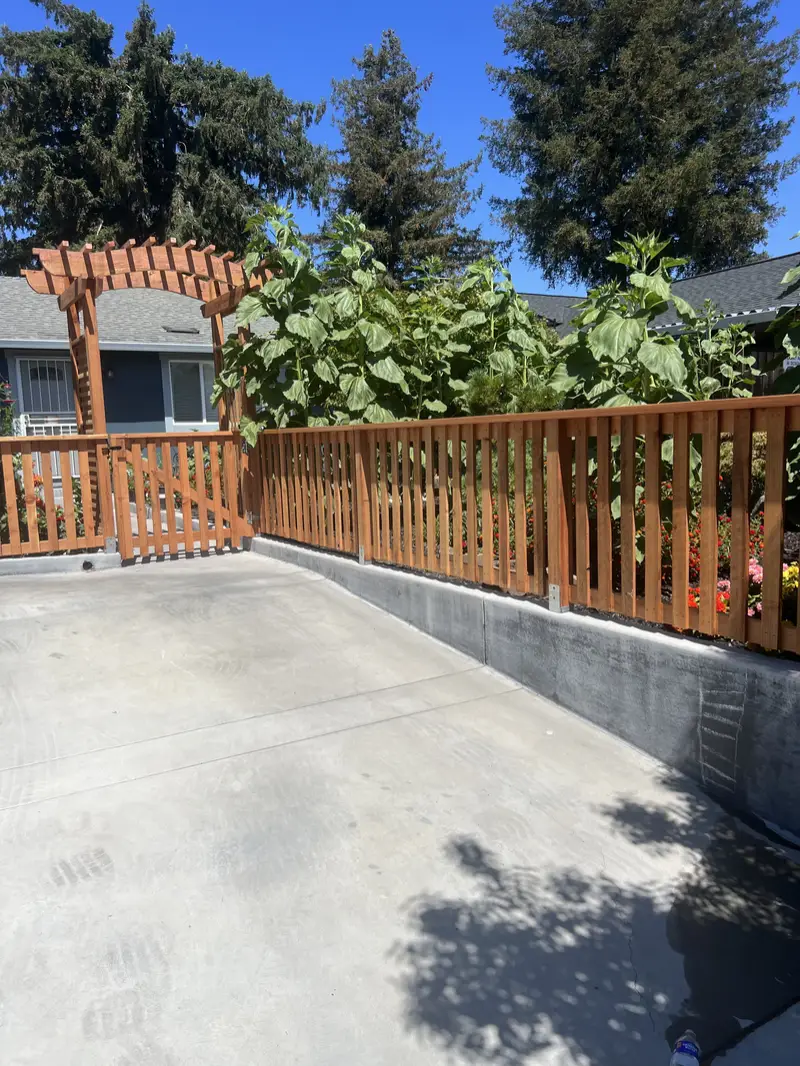 Redwood picket fence with an arbor-topped walk gate on a concrete retaining wall beside a driveway, sunflowers behind