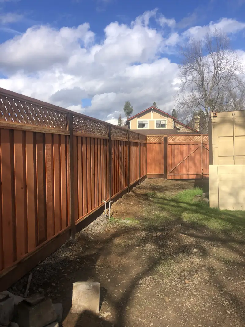 Lattice-top redwood privacy fence with a matching double gate across the back of a yard, under a cloudy blue sky