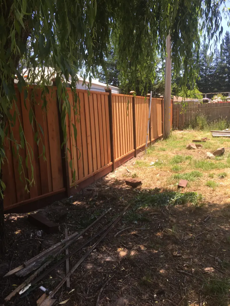 Newly built redwood board-and-batten fence with dark-stained posts and kickboard under a weeping willow, old fence boards on the ground