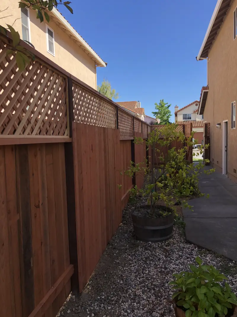 Lattice-top redwood privacy fence along a gravel side yard between two stucco houses, potted tree in front