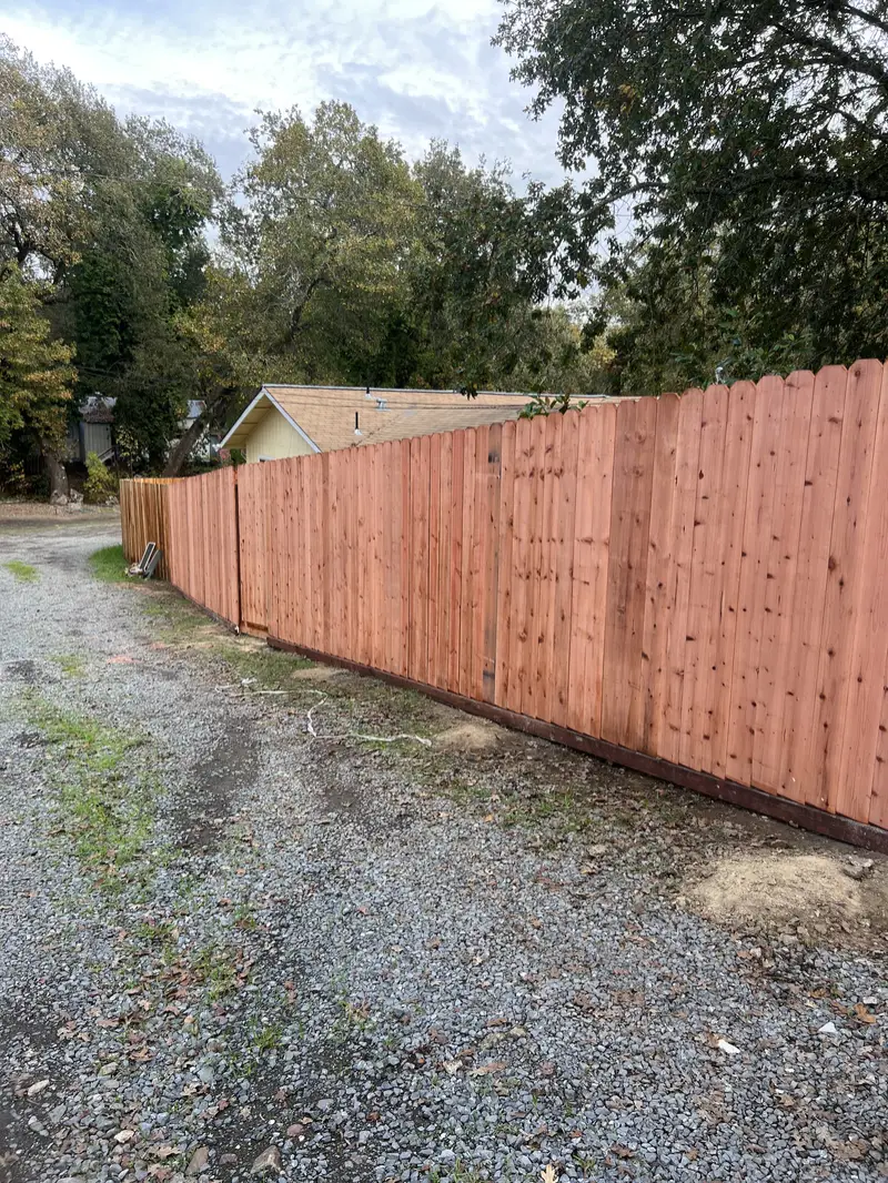 Long dog-ear redwood privacy fence along a gravel drive under oaks