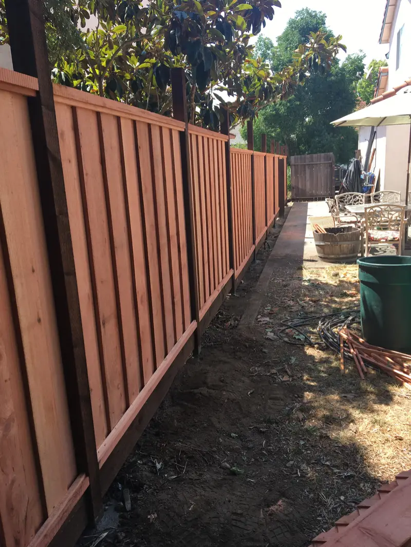 Redwood board-and-batten fence with dark-stained posts and kickboard along a side yard next to a patio set and half-barrel planter