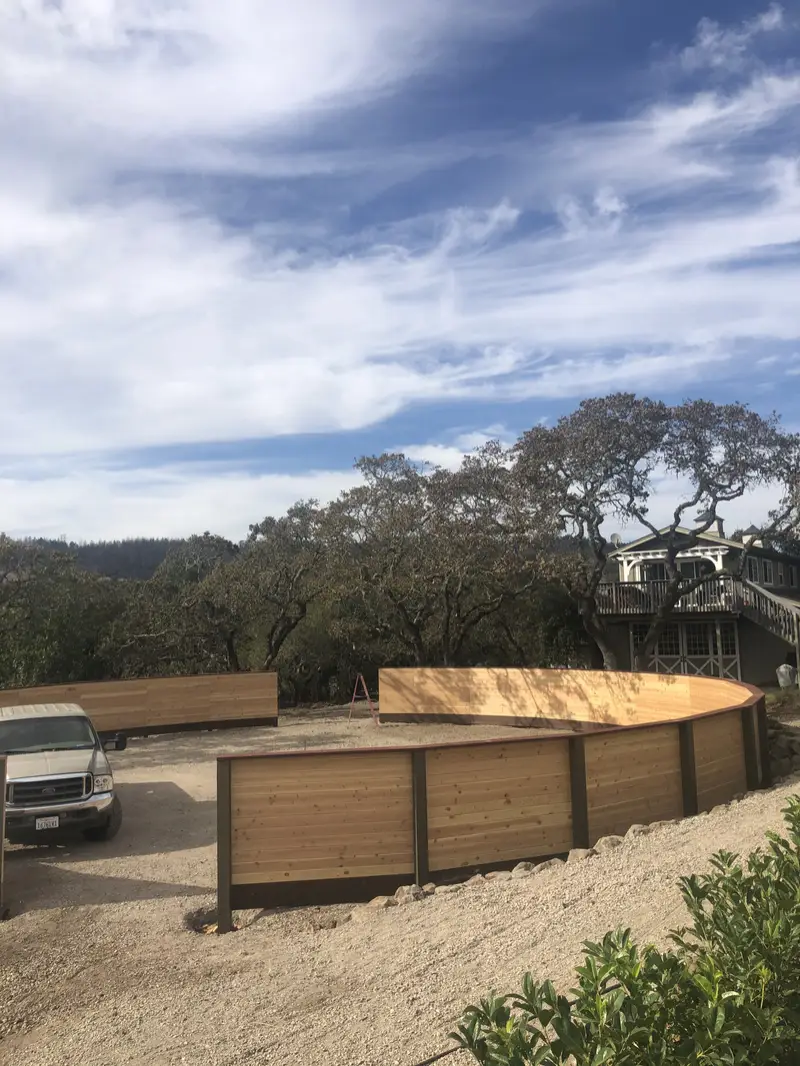 Curving solid horizontal wood round pen with dark posts on a hillside among old oaks under a streaky sky