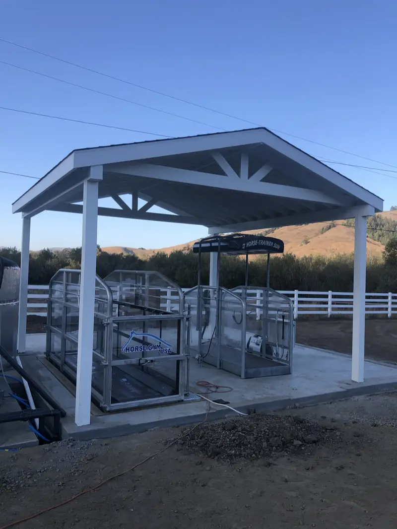 White gable-roof open pavilion sheltering horse exercise equipment on a concrete pad, white three-rail vinyl ranch fence and golden hills behind