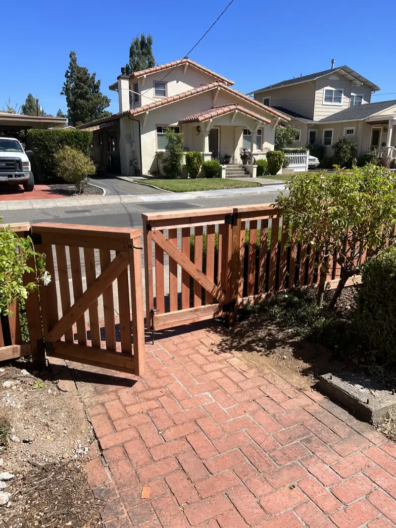 Redwood picket double walk gate with Z-braces standing open over a herringbone brick path to the street
