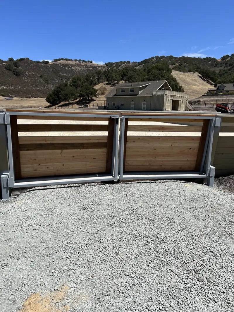 Closed double driveway gate with galvanized steel frames and horizontal redwood infill on a gravel drive, riding arena, barn and oak hills behind