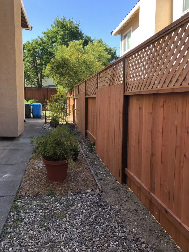 Lattice-top redwood privacy fence with dark-stained posts along a gravel side-yard planting strip beside a stucco house