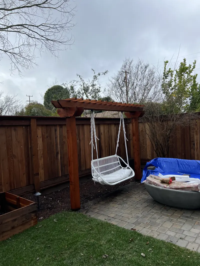 Redwood pergola-style swing arbor over a paver patio with a hanging swing chair, redwood privacy fence and raised beds behind