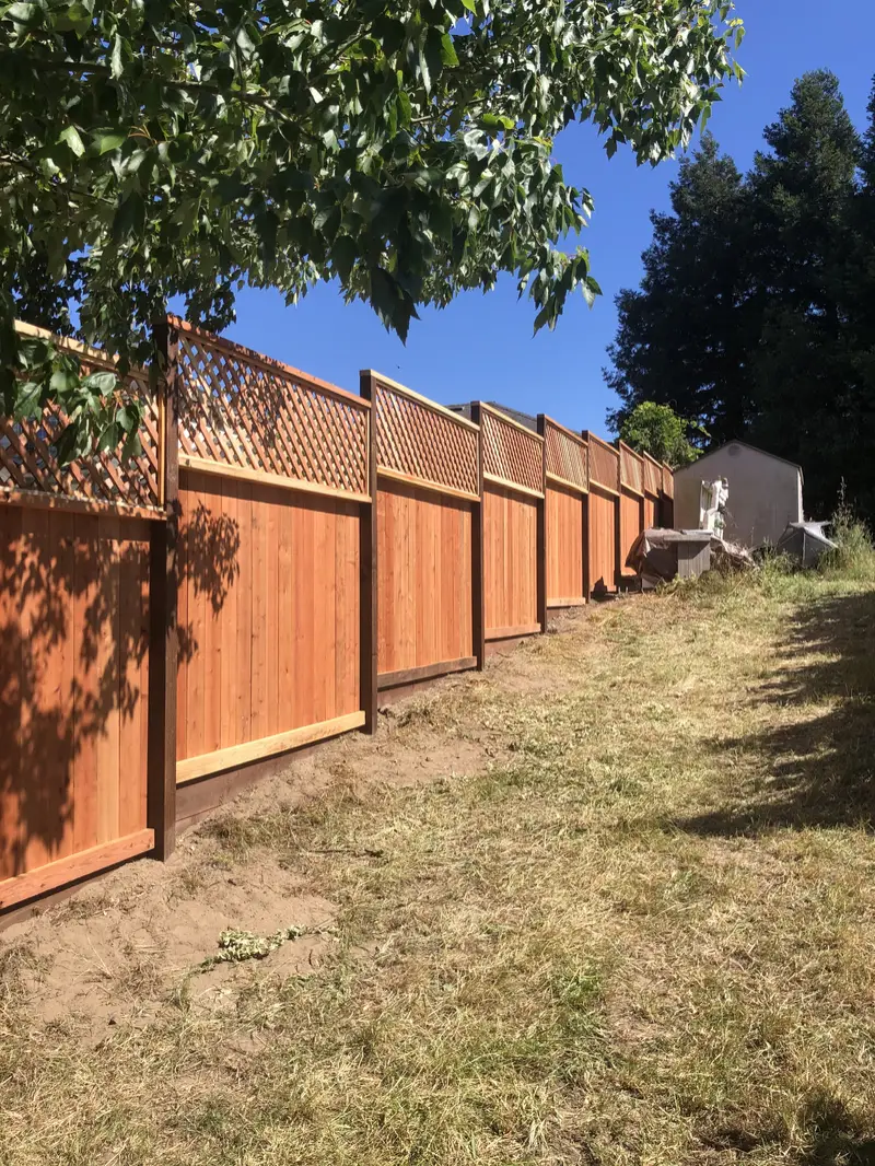 Lattice-top redwood privacy fence with dark-stained posts and kickboard along a grassy backyard under trees