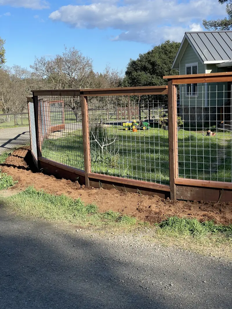 Corner of a redwood-framed welded-wire fence with kickboard beside a gravel drive, green farmhouse behind