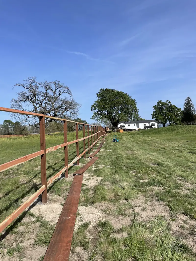 Redwood post-and-rail frame of a long pasture fence going up across a green field toward oaks
