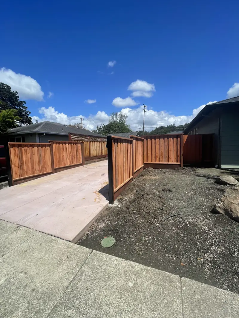 Redwood framed-panel fence with a black-framed walk gate along a new concrete driveway under puffy clouds