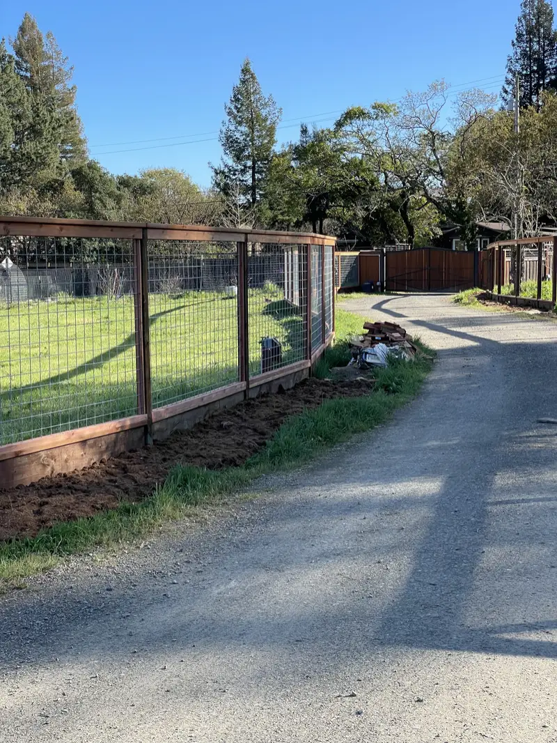 Redwood-framed welded-wire fence with kickboard along a gravel country lane leading to a wood driveway gate