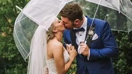 A groom in a navy tuxedo kissing his bride on their wedding day under an umbrella