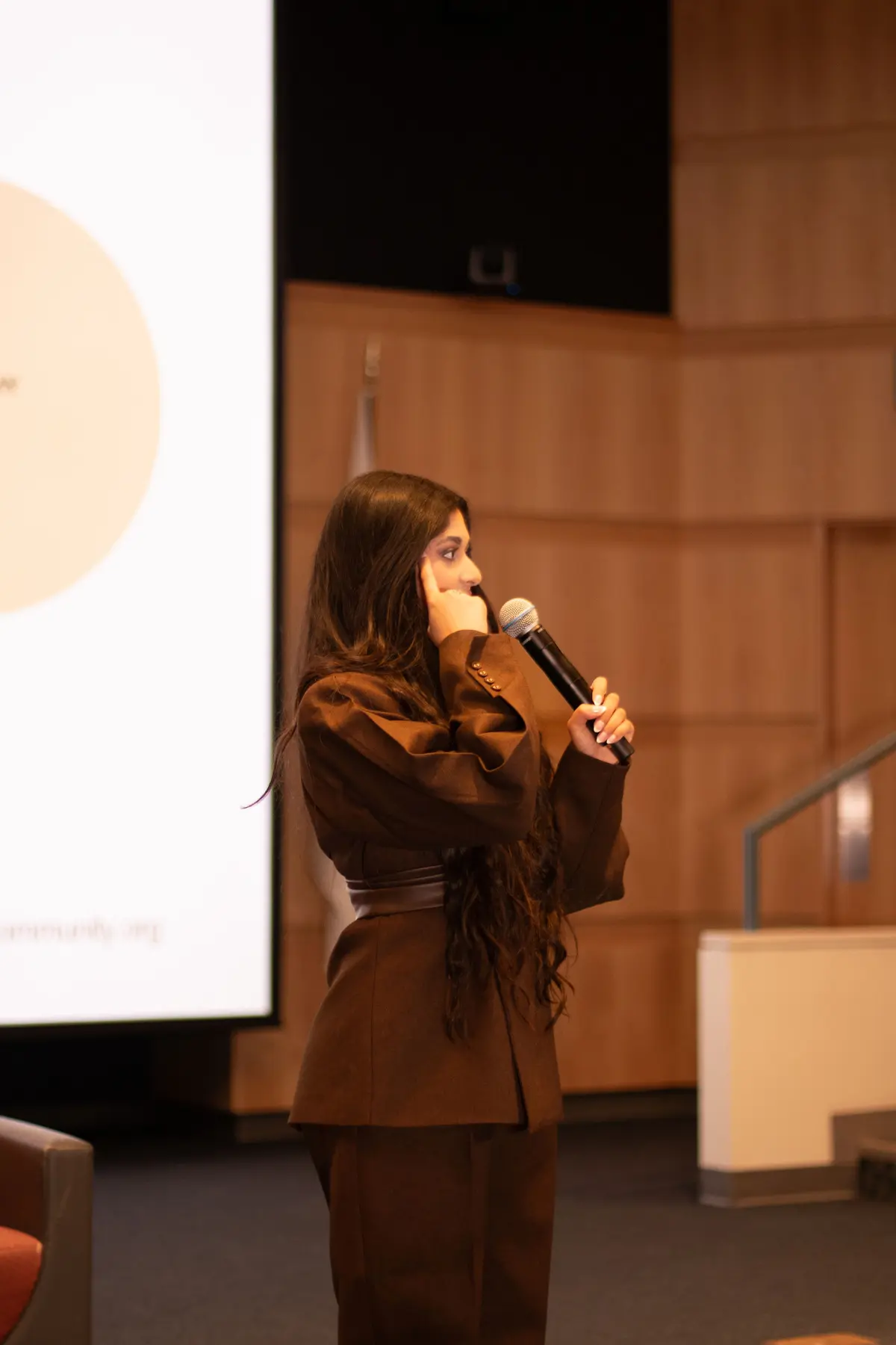 Amber Farooqui speaking with a microphone in a brown suit