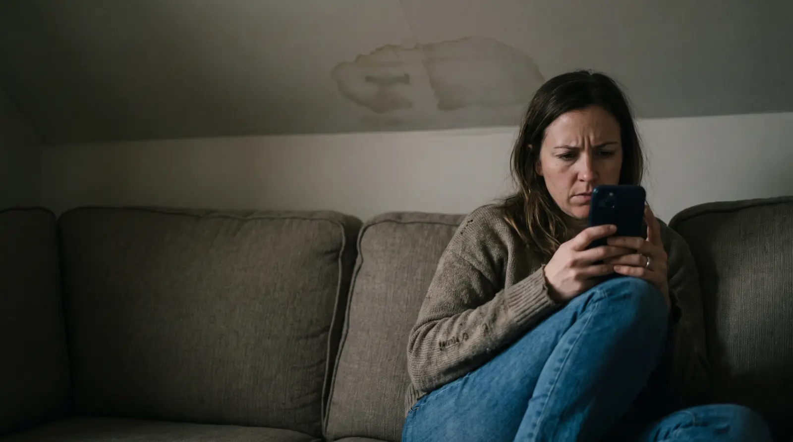 A homeowner checking her phone on the sofa at night, a damp patch visible on the ceiling.