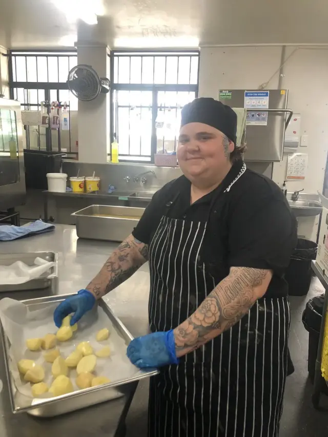 Goodness kitchen trainee in an apron and gloves preparing a tray of food