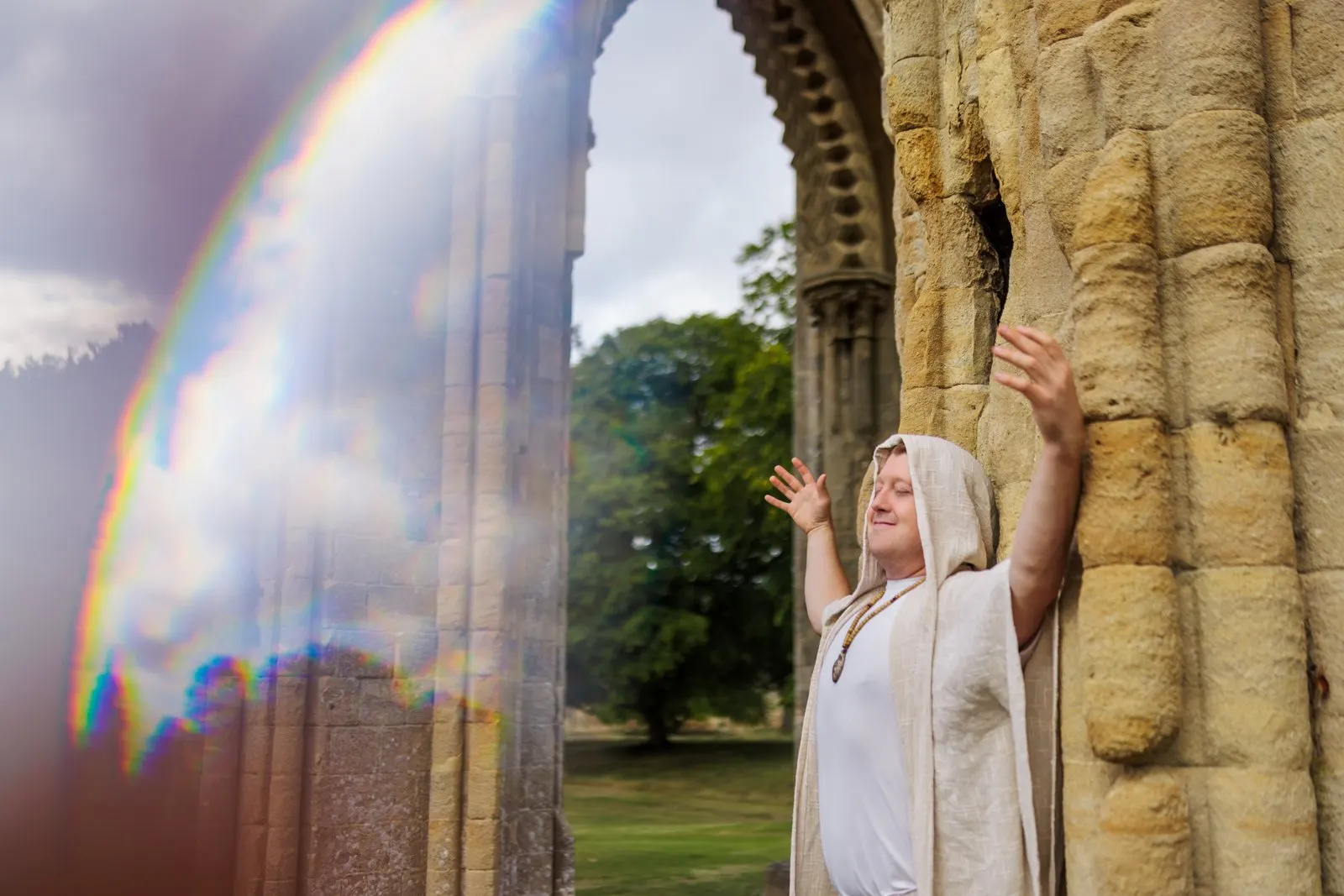 Jerome Zoran in a linen robe beneath the arches of Glastonbury Abbey, with a rainbow of light