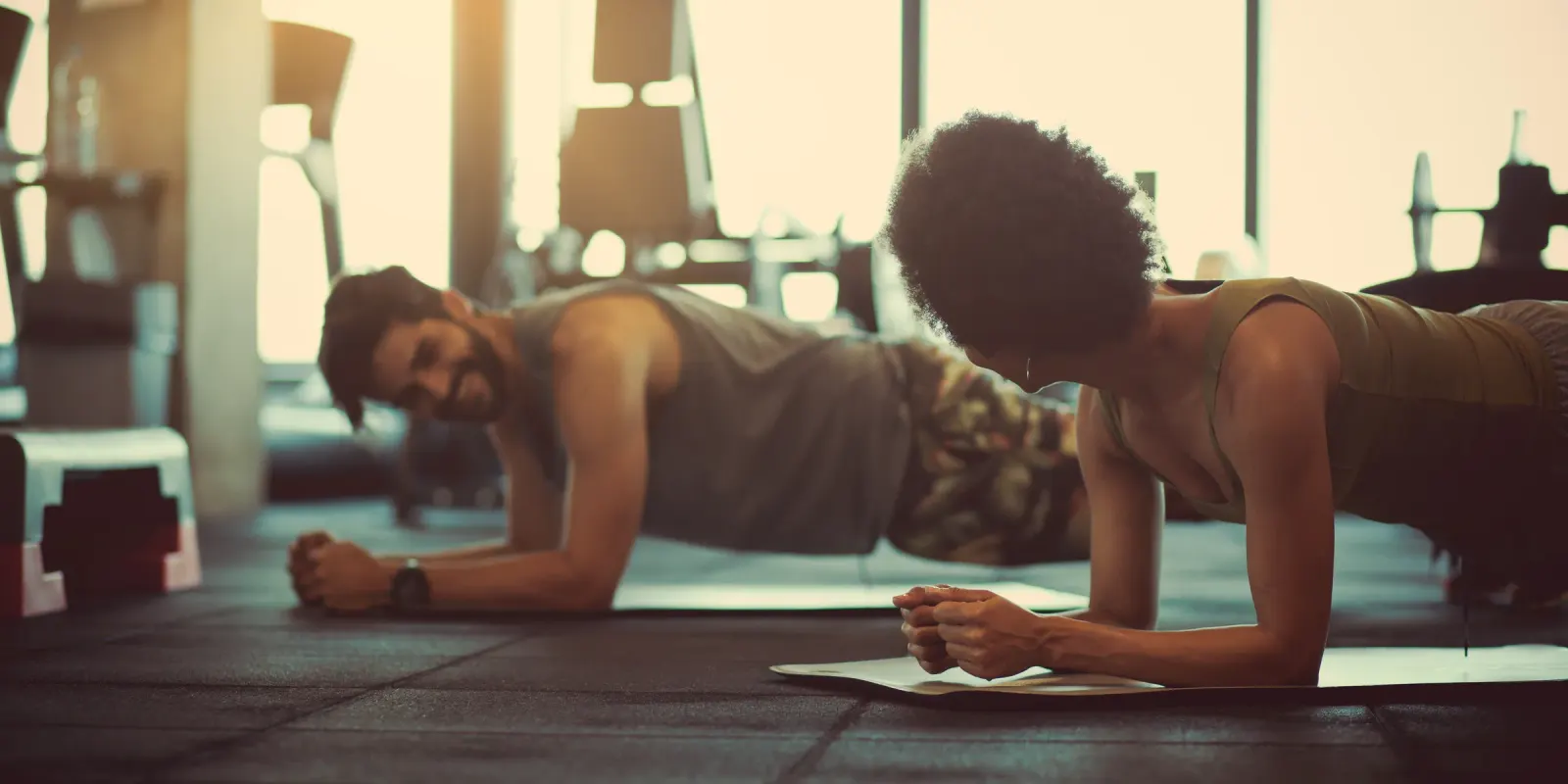 Man and woman holding a plank side by side in a sunlit gym