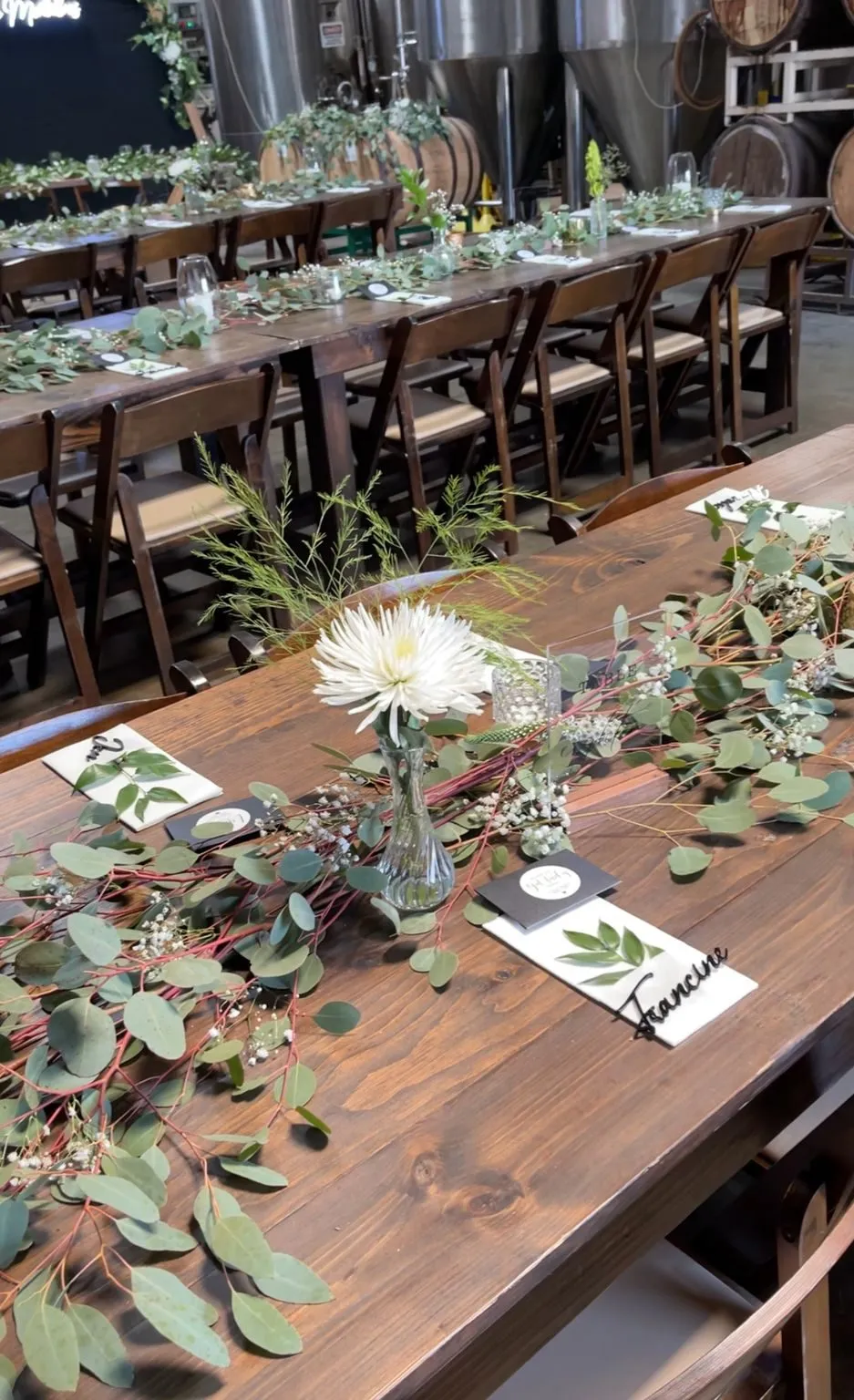 A wooden party table dressed with greenery, white flowers and place cards