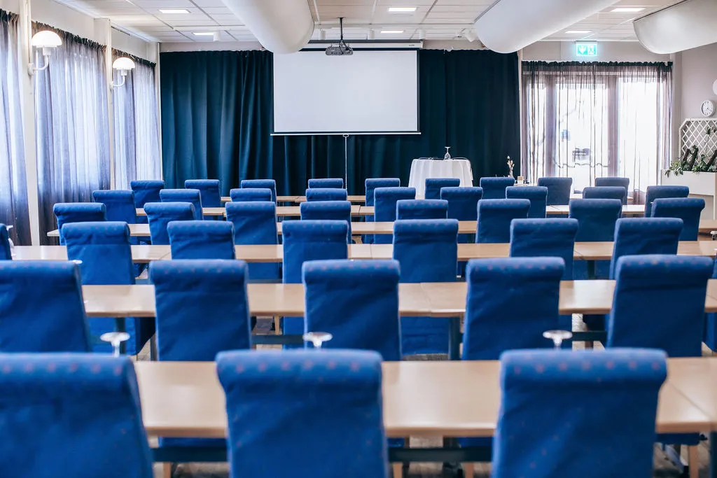 Lokalen i skolsittning med blå stolar, projektorduk och ljusa gardiner