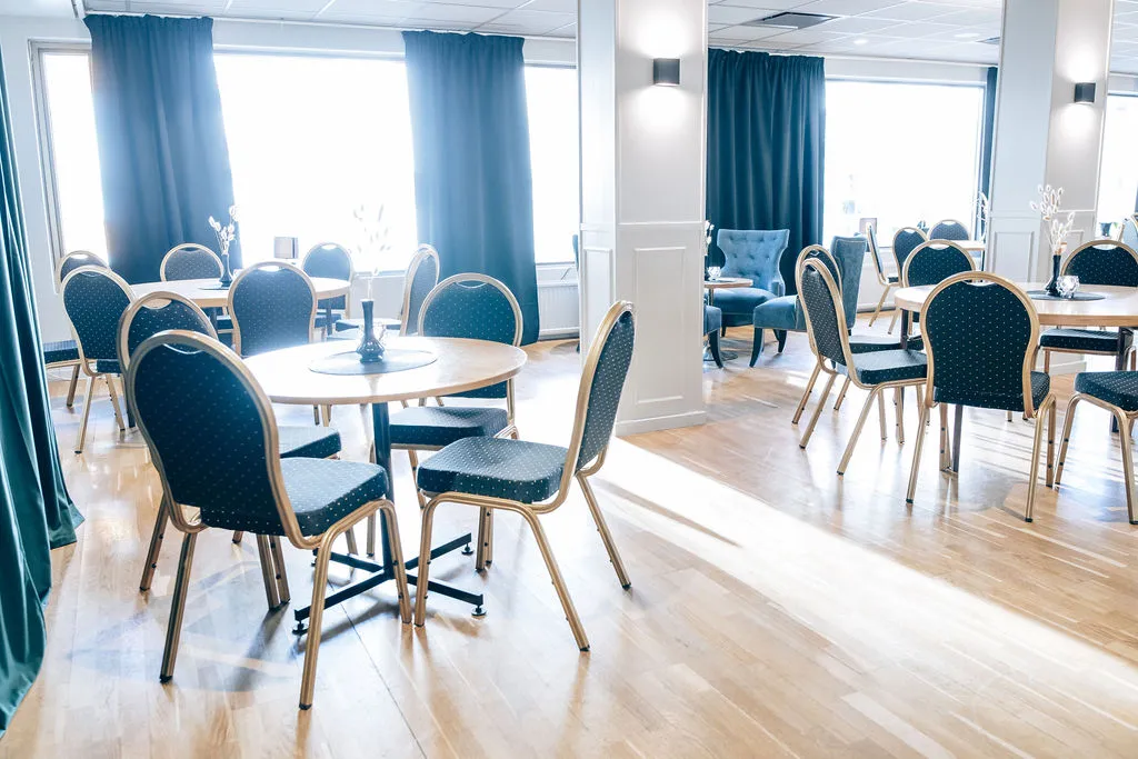 Ljus restaurang med runda bord, guldfärgade stolar och petrolblå gardiner