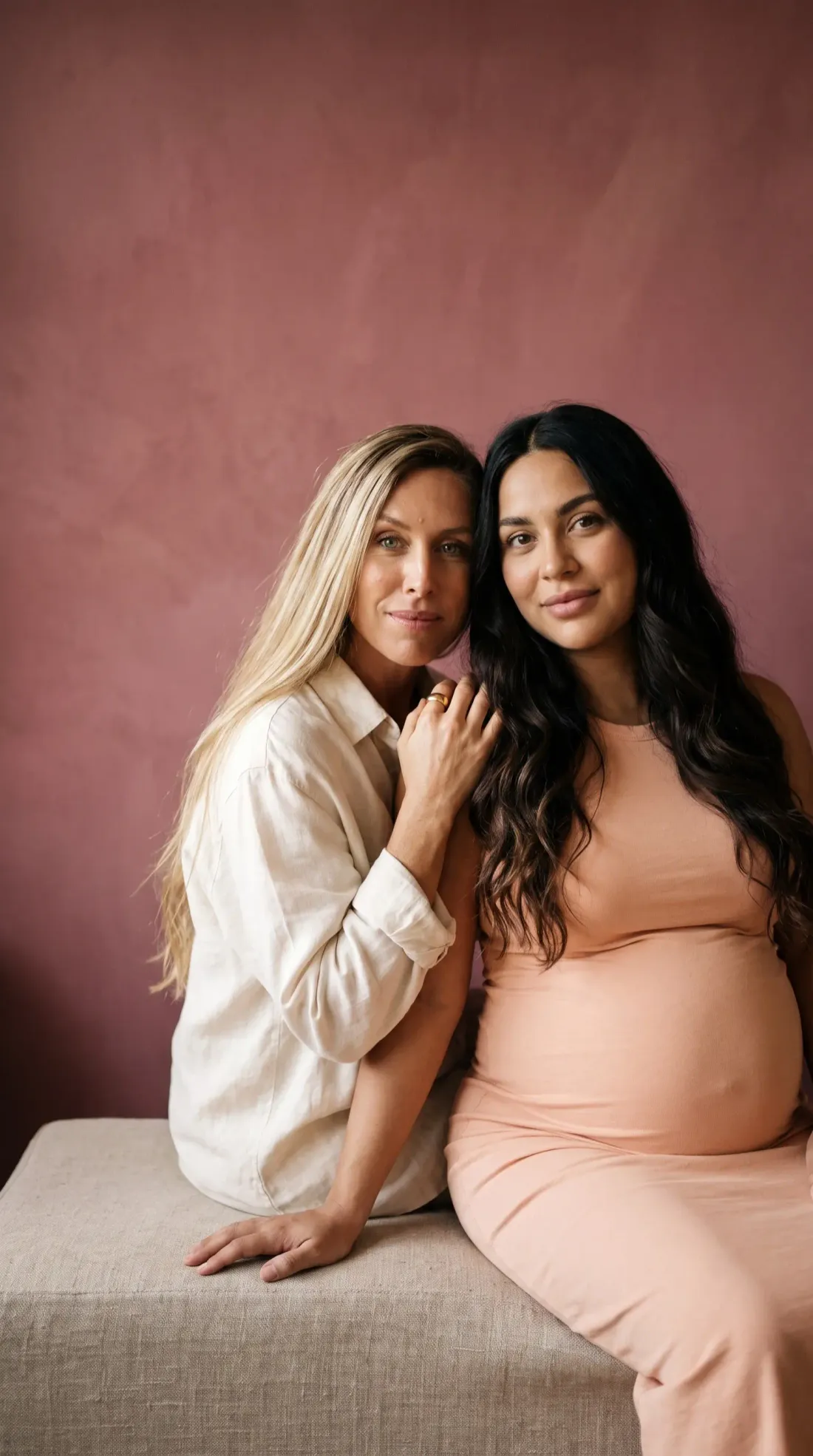 Deux femmes assises côte à côte devant un mur rose : l’une, blonde, pose la main sur l’épaule de l’autre, enceinte