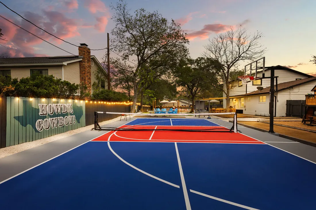 Multi-use court in blue and red with a basketball goal under string lights at dusk, Austin