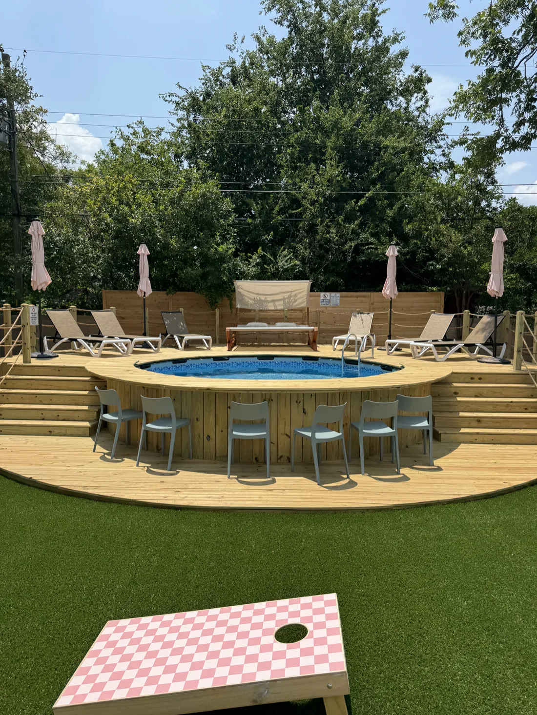 Round wood pool deck with bar seating around an above-ground pool, cornhole on the turf