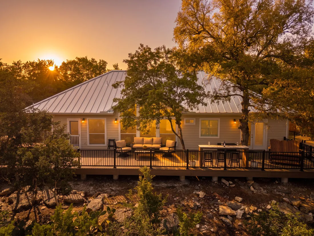 Composite deck with black metal railing across the back of a metal-roof home, lounge seating and a bar-height table at sunset