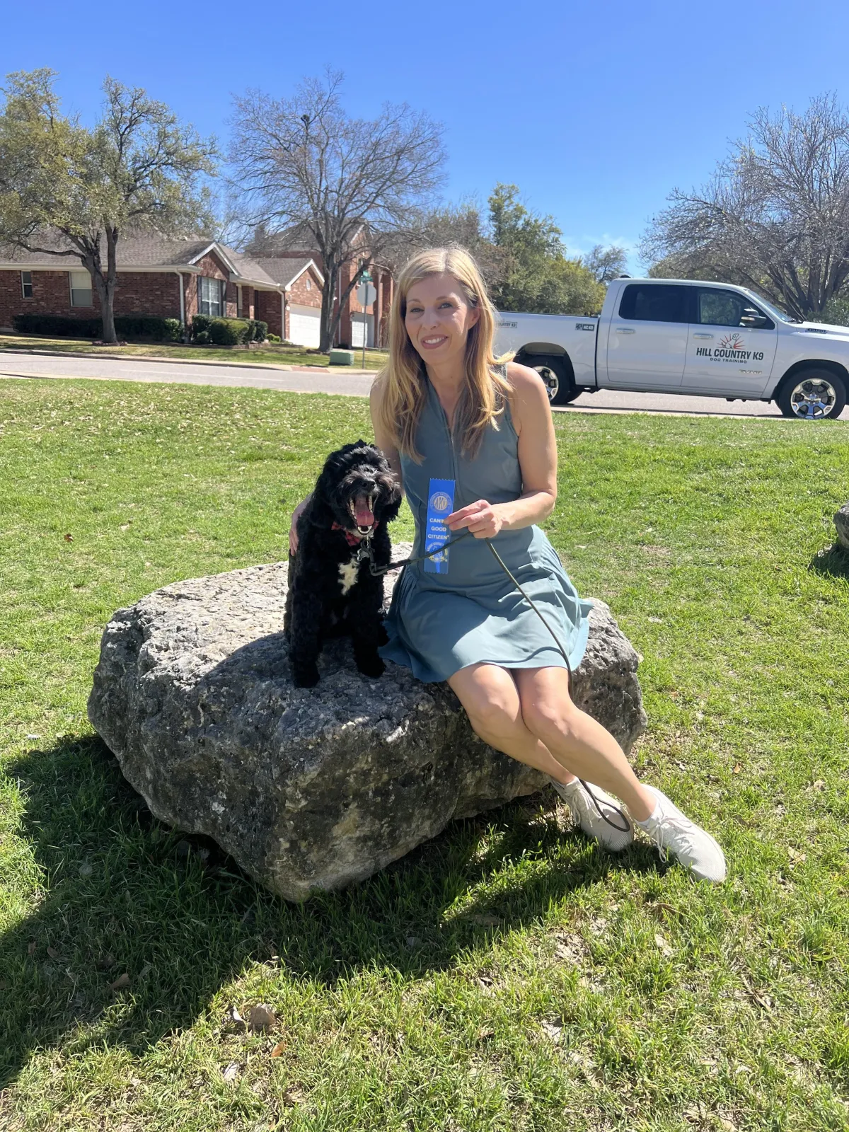 Dog owner displaying an AKC Canine Good Citizen ribbon with her certified dog and the Hill Country K9 truck behind them