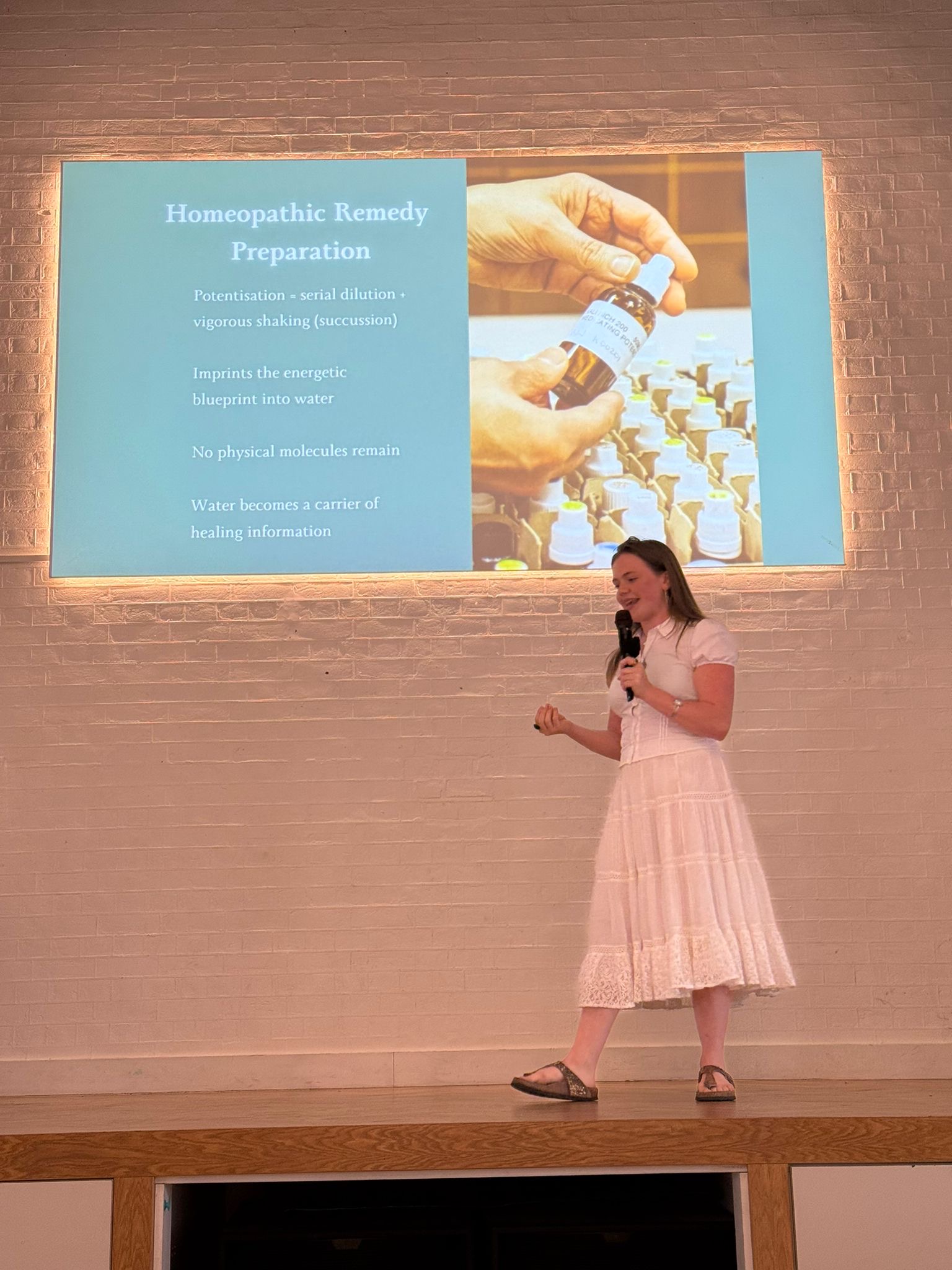 A woman in a white dress stands on a stage holding a microphone, presenting in front of a projected slide titled “Homeopathic Remedy Preparation.” The slide shows hands holding a homeopathic remedy bottle alongside text explaining potentisation, water as a carrier of healing information, and the imprinting of energetic information into water. The setting appears to be an indoor talk or lecture space with a white brick wall backdrop.